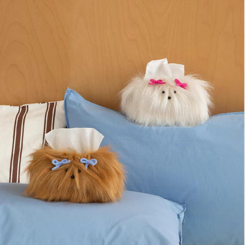 Two fluffy dog-shaped tissue holders on a bed with blue pillows and a wooden headboard.